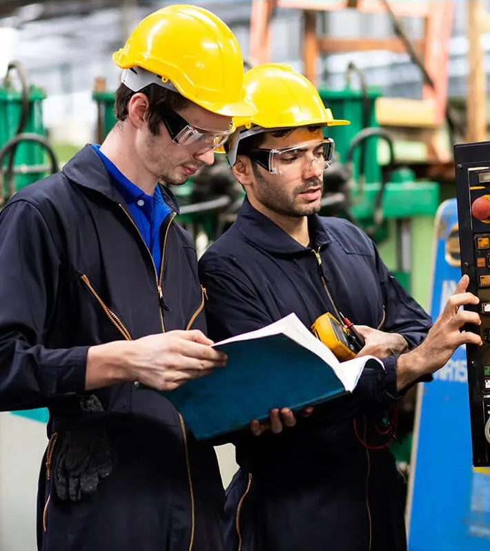 Two electricians working at an industrial factory wearing uniforms and hard hats. The electricians are performing mechanical repair and operating lathe machinery.
